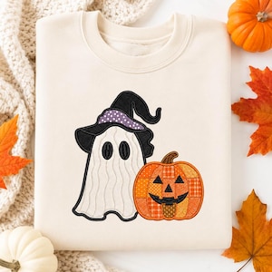 May include: A cream-colored crewneck sweatshirt featuring an embroidered design of a white ghost wearing a black witch hat with purple polka dots, positioned next to a jack-o'-lantern pumpkin with a patchwork pattern in shades of orange.