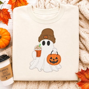 Könnte beinhalten: Ein cremefarbenes Sweatshirt mit einer Stickerei eines weißen Geistes, der eine braune Mütze trägt. Der Geist hält einen braunen Eiskaffee mit einem grünen Strohhalm in der einen Hand und trägt einen orangefarbenen Kürbiseimer in der anderen.