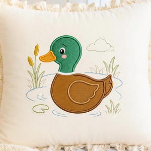 May include: Cream-colored decorative pillow with a cartoon duck embroidery. The duck has a green head, brown body, and orange beak, surrounded by embroidered cattails, grass, and a cloud. The pillow has a ruffled edge.