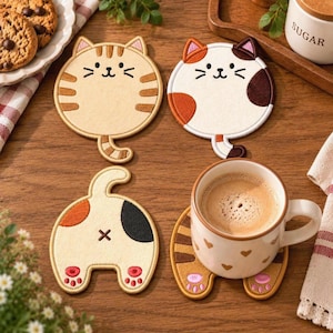 May include: Four round felt coasters featuring embroidered cat designs. Two coasters show cat faces, one in tan with stripes and one in white with orange and brown patches. Two coasters show cat backsides with pink paw pads and tails.
