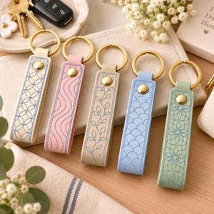 Puede incluir: Cinco llaveros rectangulares con anillas de metal dorado y botones a presión. Cada correa es de un color diferente: crema, rosa, beige, azul claro y verde salvia, decoradas con intrincados patrones bordados en azul que incluyen círculos, ondas y hojas.