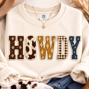May include: A cream-colored crewneck sweatshirt featuring the word HOWDY in large block letters. Each letter has a unique pattern: brown geometric, cow print, mustard cactus, brown gingham, and navy blue with gold starbursts.