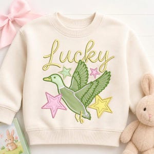 May include: A cream-colored crewneck sweatshirt featuring an embroidered design of a flying green duck. Above the duck, the word Lucky is stitched in yellow cursive script. The design includes three stars in light green, pink, and yellow colors.