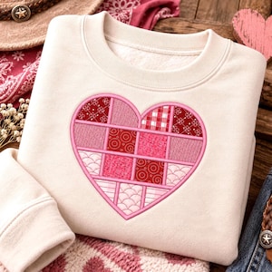 Könnte beinhalten: Ein cremefarbenes Sweatshirt mit Rundhalsausschnitt und einem großen, zentralen Herzmotiv. Das Herz ist in einem Patchwork-Gittermuster gestaltet und zeigt verschiedene Rosa- und Rottöne mit unterschiedlichen geometrischen, floralen und strukturierten Drucken.