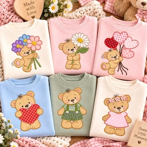 May include: Six crewneck sweatshirts in cream, pink, blue, and sage green, each featuring an embroidered teddy bear. The bears are depicted holding flowers, heart-shaped balloons, a large red heart, wearing green overalls, or wearing a pink dress.
