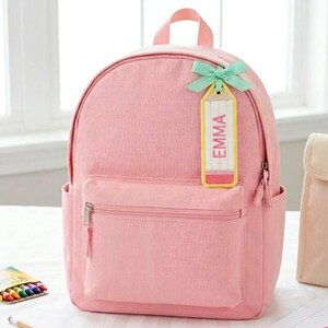 Pode incluir: Uma mochila rosa claro com um bolso frontal com z&iacute;per e dois bolsos laterais. Uma etiqueta amarela em forma de l&aacute;pis com o nome "EMMA" &eacute; presa com um la&ccedil;o verde claro. A mochila tem uma al&ccedil;a superior e um fecho de z&iacute;per.