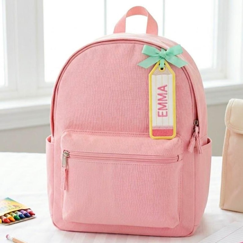 Pode incluir: Uma mochila rosa claro com um bolso frontal com z&iacute;per e dois bolsos laterais. Uma etiqueta amarela em forma de l&aacute;pis com o nome "EMMA" &eacute; presa com um la&ccedil;o verde claro. A mochila tem uma al&ccedil;a superior e um fecho de z&iacute;per.