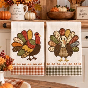 May include: Two off-white waffle-weave kitchen towels featuring embroidered turkey designs. The left towel has a turkey with colorful patterned feathers and a red wattle, while the right towel features a turkey with brown and floral feathers.