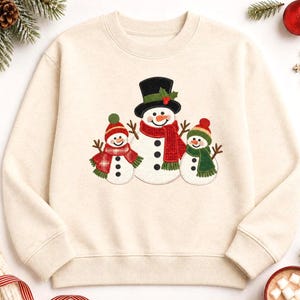 May include: A cream-coloured crewneck sweatshirt featuring a central graphic of three snowmen. The largest snowman wears a black top hat with holly, while the two smaller snowmen wear colourful knit hats and scarves in red, green, and plaid patterns.
