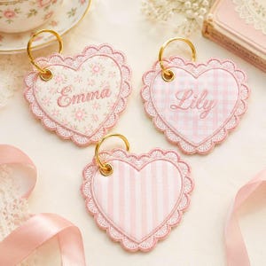 May include: Three heart-shaped keychains with gold rings. Each keychain has a pink scalloped border and a different interior design: floral, gingham, and striped. The names "Emma" and "Lily" are embroidered on two of the keychains.