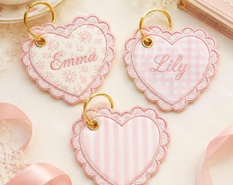 Pode incluir: Três chaveiros em forma de coração com argolas douradas. Cada chaveiro tem uma borda rosa com vieiras e um design interior diferente: floral, xadrez e listrado. Os nomes "Emma" e "Lily" são bordados em dois dos chaveiros.