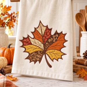 May include: A cream-colored kitchen towel featuring a large, central embroidered maple leaf design. The leaf is composed of various fabric segments in shades of orange, yellow, brown, and cream, each displaying distinct floral and dotted patterns.