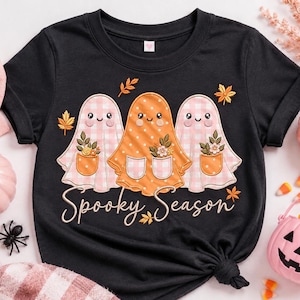 以下が含まれることがあります： 黒の半袖Tシャツで、花が入ったポケットを持つ3体の幽霊のグラフィックが描かれています。2体の幽霊はピンクのギンガムチェック柄で、中央の幽霊は白の水玉模様が入ったオレンジ色です。下にSpooky Seasonという文字がプリントされています。