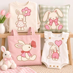 Pode incluir: Uma coleção de itens com desenhos de coelhos bordados: uma camisola creme, uma almofada axadrezada verde e branca, um saco de pano rosa, um macacão branco com folhos e um coelho de pelúcia. Os itens exibem vários motivos de coelhos.