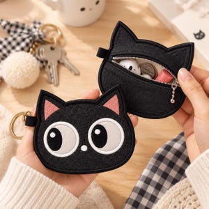 Könnte beinhalten: Zwei schwarze, katzenförmige Münztaschen aus Filz mit gestickten Details. Eine Tasche zeigt ein Gesicht mit großen weißen Augen und rosa Ohren, während die andere mit einem silbernen Reißverschluss geöffnet ist und Kleinteile wie Münzen enthält.