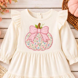 May include: A cream-coloured long-sleeve dress featuring ruffled shoulders and a gathered skirt. The centre of the bodice displays an embroidered pumpkin applique with a pink and white gingham bow and a floral pattern in shades of pink and blue.