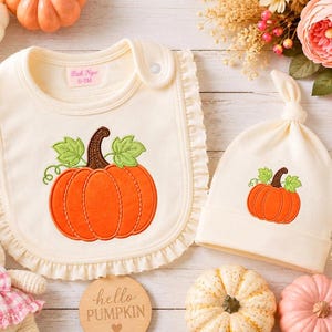 May include: A cream-coloured baby bib with a ruffled edge and a matching knotted hat, both featuring an embroidered orange pumpkin with green leaves. The bib has a label reading Bich Ngoc 0-6M and a snap closure. The set is displayed on a light surface.