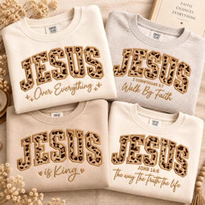 May include: Four crewneck sweatshirts in cream and light gray featuring the word JESUS in leopard print block letters. The shirts include embroidered phrases: Over Everything, Walk By Faith, is King, and The way The Truth The life.