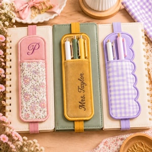 May include: Three vertical notebooks with elastic pen holders. The left is pink with a floral pattern and letter P. The center is green with a mustard yellow holder embroidered with Mrs. Taylor. The right is white with a purple gingham scalloped holder.