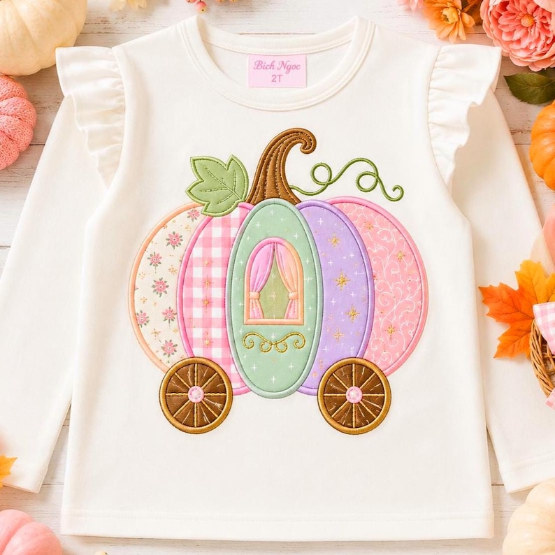 May include: A white long-sleeved shirt with ruffled shoulders and a size 2T label. The front features an embroidered pumpkin carriage design in pastel pink, purple, and green with a brown stem, green leaf, and two brown wheels with pink centres.
