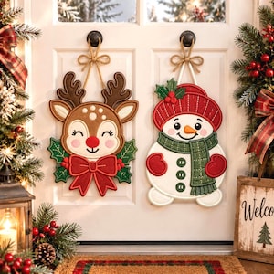 May include: Two hanging holiday decorations on a white door. One is a brown reindeer with a red nose, green holly leaves, and a large red bow. The other is a white snowman wearing a red hat, a green scarf, and red mittens, both with twine.