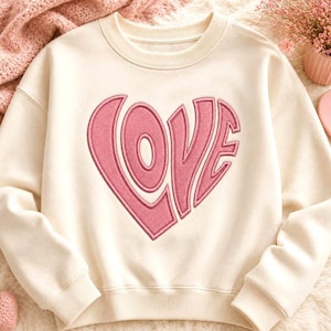 Puede incluir: Una sudadera de cuello redondo color crema con una gran aplicación rosa texturizada en forma de corazón. La palabra LOVE está integrada en el diseño del corazón con una tipografía audaz y estilizada. La prenda tiene mangas largas con puños acanalados.