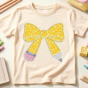 May include: A light beige t-shirt featuring a yellow bow and pencil design. The bow is filled with a white floral pattern. The pencil has a pink eraser and a gray lead. The shirt is a children's apparel item.
