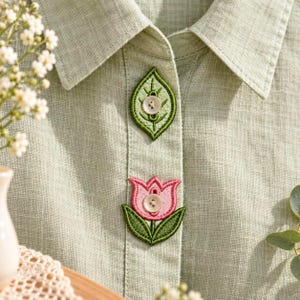 May include: A light green button-down shirt featuring two embroidered patches on the placket. The top patch is a green leaf with a white button, and the bottom patch is a pink tulip with green leaves and a white button in the center.