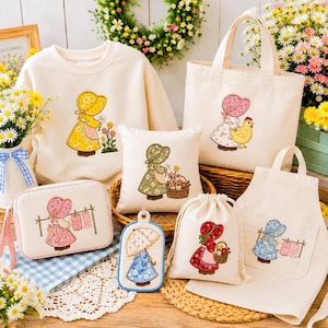 Puede incluir: Una colección de artículos textiles de color crema con figuras bordadas con capotas, que incluye una sudadera, una bolsa de tela, un cojín cuadrado, un estuche con cremallera, una etiqueta rectangular pequeña, una bolsa con cordón y un delantal.