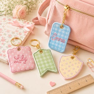 Op de afbeelding: Een verzameling gepersonaliseerde sleutelhangers in verschillende vormen en pastelkleuren. De sleutelhangers hebben geborduurde namen en patronen, waaronder ruitjes en bloemen. Goudkleurige hardware en een roze rugzak zijn ook zichtbaar.