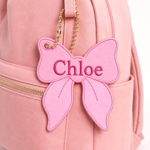 May include: A light pink backpack featuring a matching light pink bow-shaped luggage tag attached by a gold-toned ball chain. The bow tag is embroidered with the name Chloe in a dark pink thread, centered on the front of the backpack.