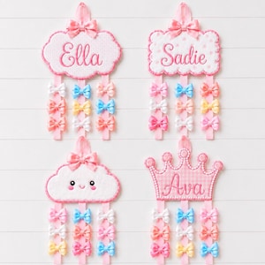 以下が含まれることがあります： Ella、Sadie、Avaの名前が刺繍されたものや、笑顔の雲が描かれたものなど、4つの布製ウォールオーガナイザー。各ピースの上部にはリボンがあり、3本の垂直なリボンにはピンク、青、黄色、白の小さなカラフルなリボンが飾られています。