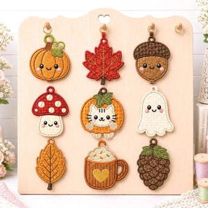May include: A set of nine embroidered hanging ornaments on a light wood display board. The designs include a pumpkin, maple leaf, acorn, mushroom, cat in a pumpkin, ghost, leaf, coffee mug with a heart, and pinecone, all featuring cute faces.