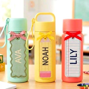 May include: Three colorful water bottles with personalized name tags. The bottles are mint green, yellow, and coral. Each bottle has a ribbon and a tag with a name: AVA, NOAH, and LILY. The yellow bottle is designed to look like a pencil.