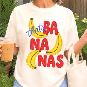 Puede incluir: Una camiseta de manga corta color crema con un gráfico bordado de tres plátanos amarillos y el texto rojo "BA NA NAS", junto con la palabra "that's" en cursiva azul situada sobre los plátanos, creando un diseño divertido y colorido.