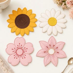 May include: A set of four flower-shaped coasters featuring embroidered designs. One is a yellow sunflower with a brown centre, one is a white daisy with a yellow centre, and two are pink blossoms with detailed stitching and central circular patterns.