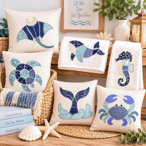 May include: A collection of cream-coloured throw pillows and towels featuring blue and teal marine life appliques, including a manta ray, sea turtle, whale, seahorse, whale tail, and crab, arranged with books and a starfish on a woven surface.