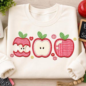 May include: A cream-colored crewneck sweatshirt featuring an embroidered design of three apples in various states: a bitten apple core, a sliced apple half, and a whole apple. The apples feature red gingham fabric patterns and green leaves.
