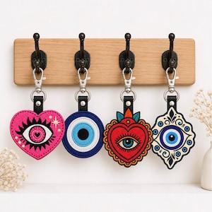 May include: Four embroidered keychains hanging from a light wood wall rack with black metal hooks. The keychains feature a pink heart with an eye, a blue circular evil eye, a red heart with an eye, and a decorative blue and white evil eye shape.