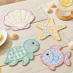 Puede incluir: Cuatro posavasos decorativos de tela con forma de concha, estrella de mar, tortuga y pez. La concha es blanca con un estampado de margaritas y un borde lila. La estrella de mar tiene un estampado de cuadros.