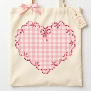 May include: Cream-colored tote bag with a large pink gingham heart design. The heart is outlined with a scalloped edge and adorned with small bows. A pink satin bow is tied at the top, and a tag with text hangs from the handle.