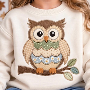 Puede incluir: Una sudadera de color crema con un gran diseño de búho bordado en el centro. El búho está compuesto por varios parches de tela en marrón, beige, verde y azul, con patrones que incluyen cuadros vichy, lunares y margaritas blancas en una rama.