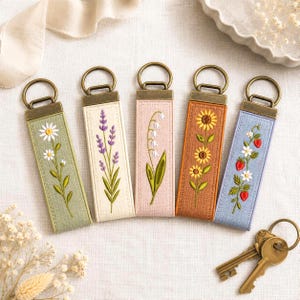 Pode incluir: Cinco porta-chaves retangulares de tecido com ferragens de metal cor de bronze. Cada um apresenta desenhos botânicos bordados: margaridas brancas em verde sálvia, lavanda roxa em creme, lírios do vale em rosa, girassóis em bege e morangos em azul.