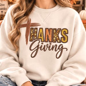 May include: A cream-colored crewneck sweatshirt featuring a brown graphic design that reads "THANKS" in block letters with a plaid and floral pattern, and "Giving" in brown cursive script, accented by a brown cross-shaped brushstroke graphic.