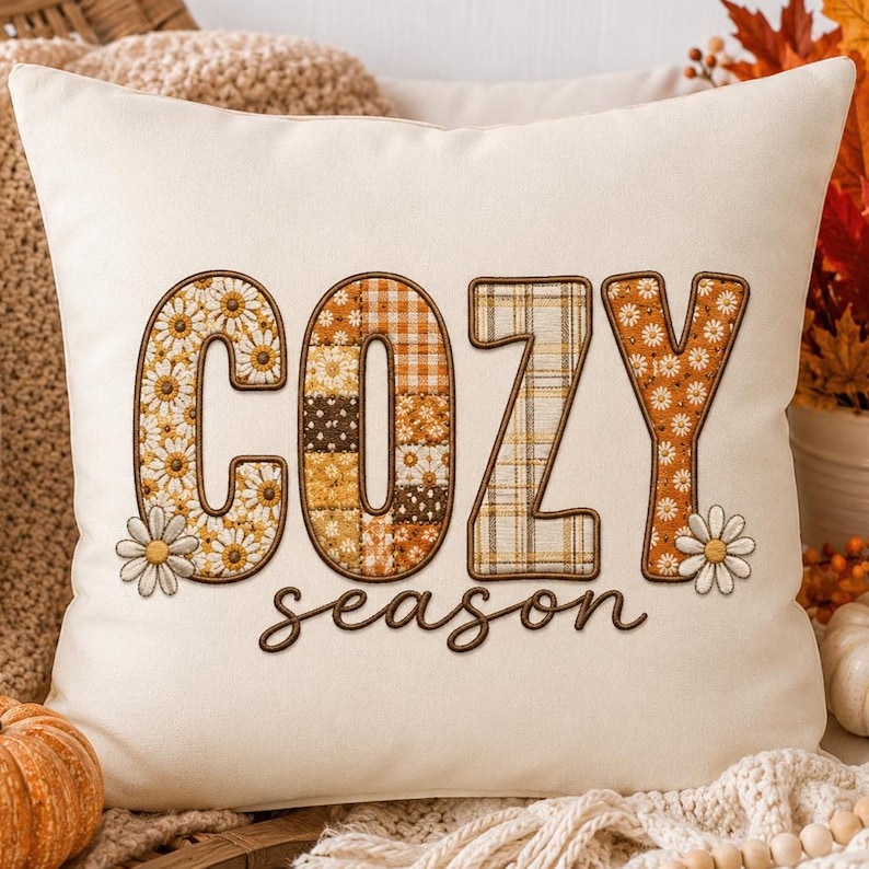 May include: A square off-white throw pillow featuring the word COZY in large, block-style letters filled with various autumn-themed patterns including daisies, plaid, and checkered prints. The word season is written below in brown script.