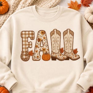 May include: A cream-colored crewneck sweatshirt featuring the word FALL in large letters. The F is brown gingham with a horseshoe, the A is floral with a pumpkin, and the two Ls are shaped like western cowboy boots with floral accents.