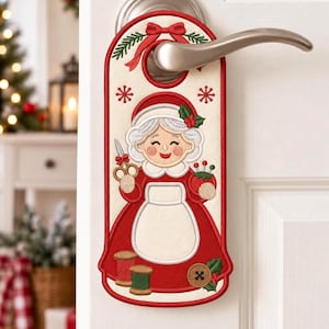 May include: A cream-colored fabric door hanger featuring an embroidered design of a smiling character with white hair wearing a red dress and white apron. The character holds scissors and a pincushion, with spools of thread and a button below.