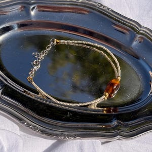 Peut inclure: Un bracelet délicat composé de deux rangées de petites perles argentées, de perles dorées et d'une perle centrale ovale ambrée. Le bracelet est présenté sur un plateau argenté.
