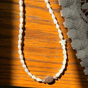 Peut inclure: Un collier composé d'un rang de petites perles blanches en forme de goutte avec une finition nacrée. Le collier comprend une perle en forme d'étoile à cinq branches, deux petites perles intercalaires transparentes et une breloque ovale centrale.