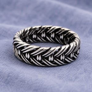 Puede incluir: Un anillo plateado con un diseño tejido. El anillo tiene una superficie texturizada y una apariencia de banda. El anillo está sobre un fondo azul.
